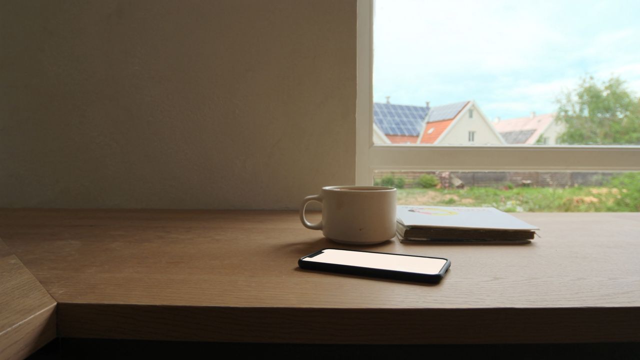 Handy und Kaffeetasse auf einem Holztisch am Fenster, draußen Häuser mit Solarmodulen auf den Dächern